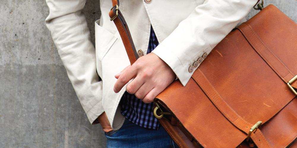Close-up of person wearing beige blazer and checkered shirt holding brown genuine leather laptop bag over shoulder against gray wall